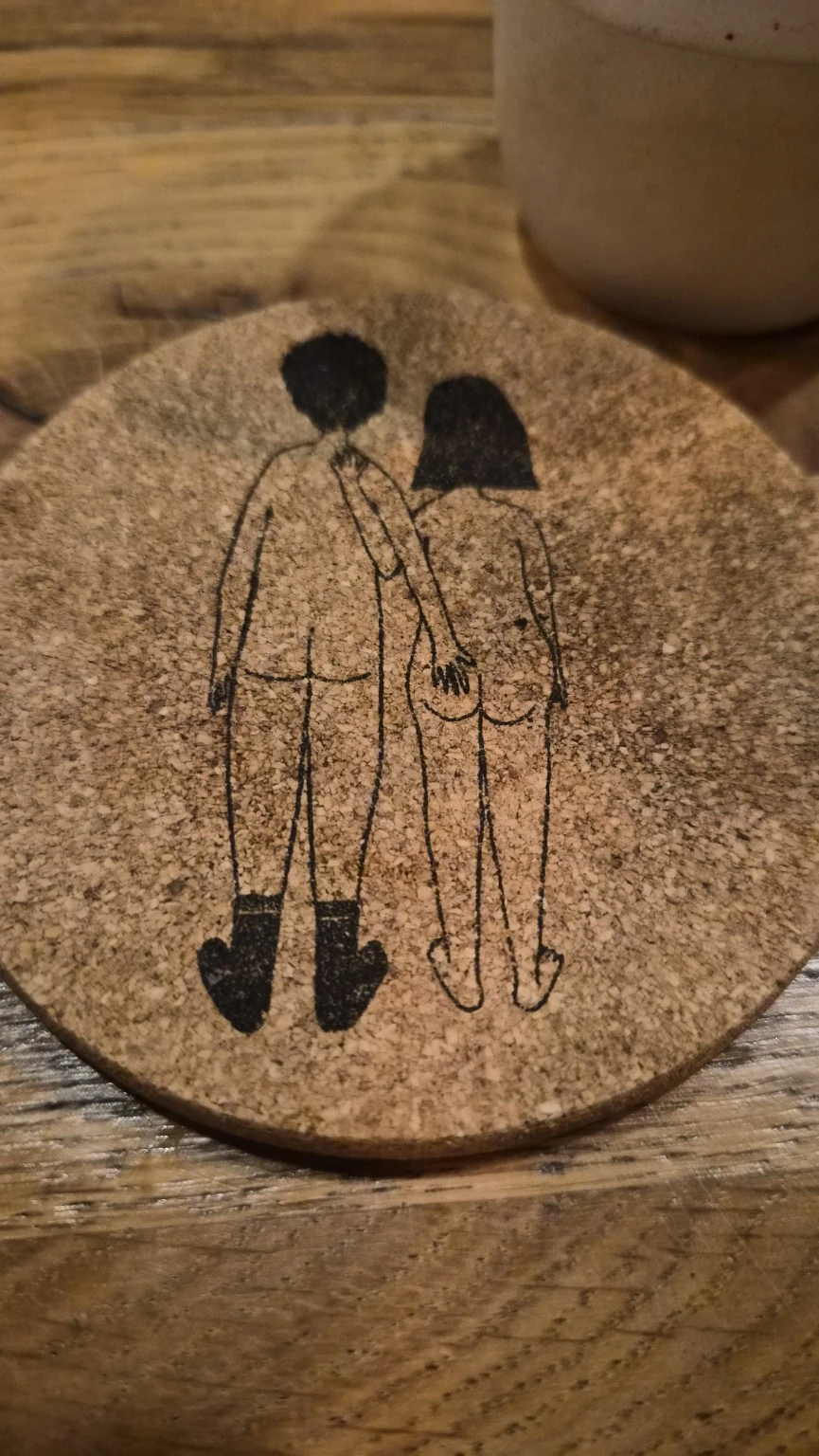 Korkuntersetzer mit gezeichnetem Paar und einer Hand am Po auf dem Holztisch im Fackelgarten Plau am See.