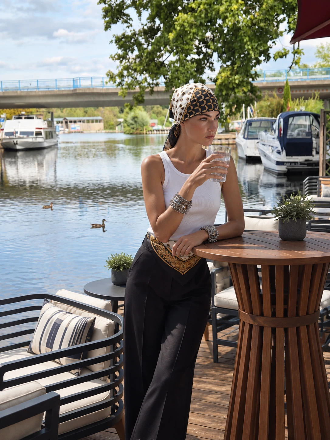 Frau mit gemustertem Kopftuch an der Uferterrasse des Fackelgartens, ein Glas Wasser in der Hand, Boote und Brücke im Hintergrund