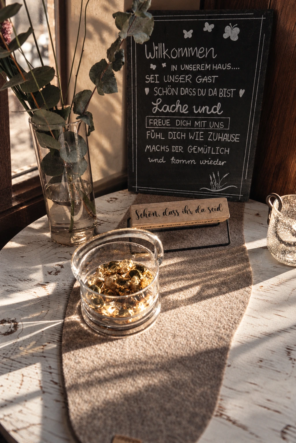 Willkommensschild, Eukalyptus und Schokoladen-Schälchen auf einem sonnigen Tisch in Marias KostBar