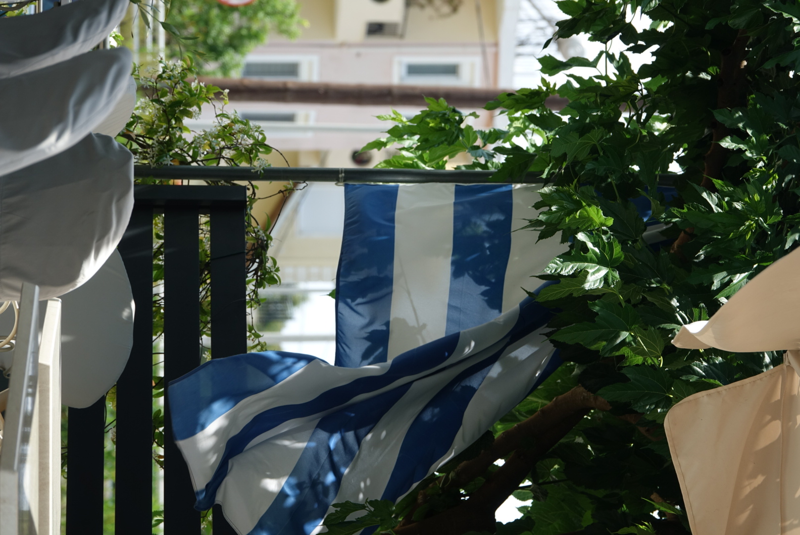 Griechische Flagge im Wind zwischen grünen Blättern und weißen Stühlen.