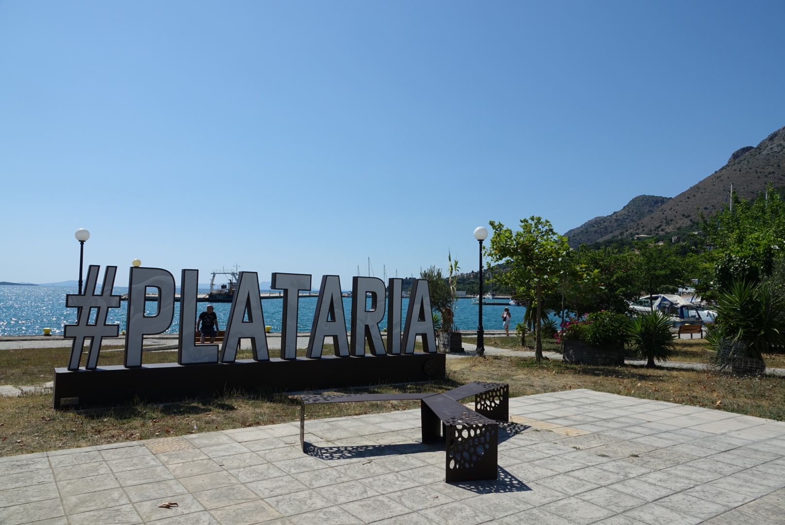 Das große Plataria-Schild am Hafen mit Meer und Bergen im Hintergrund.